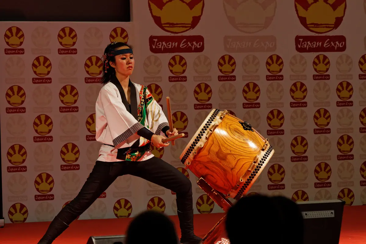 A drummer in a white happi coat and headband lunges low beside a lacquered taiko, a stick in each hand