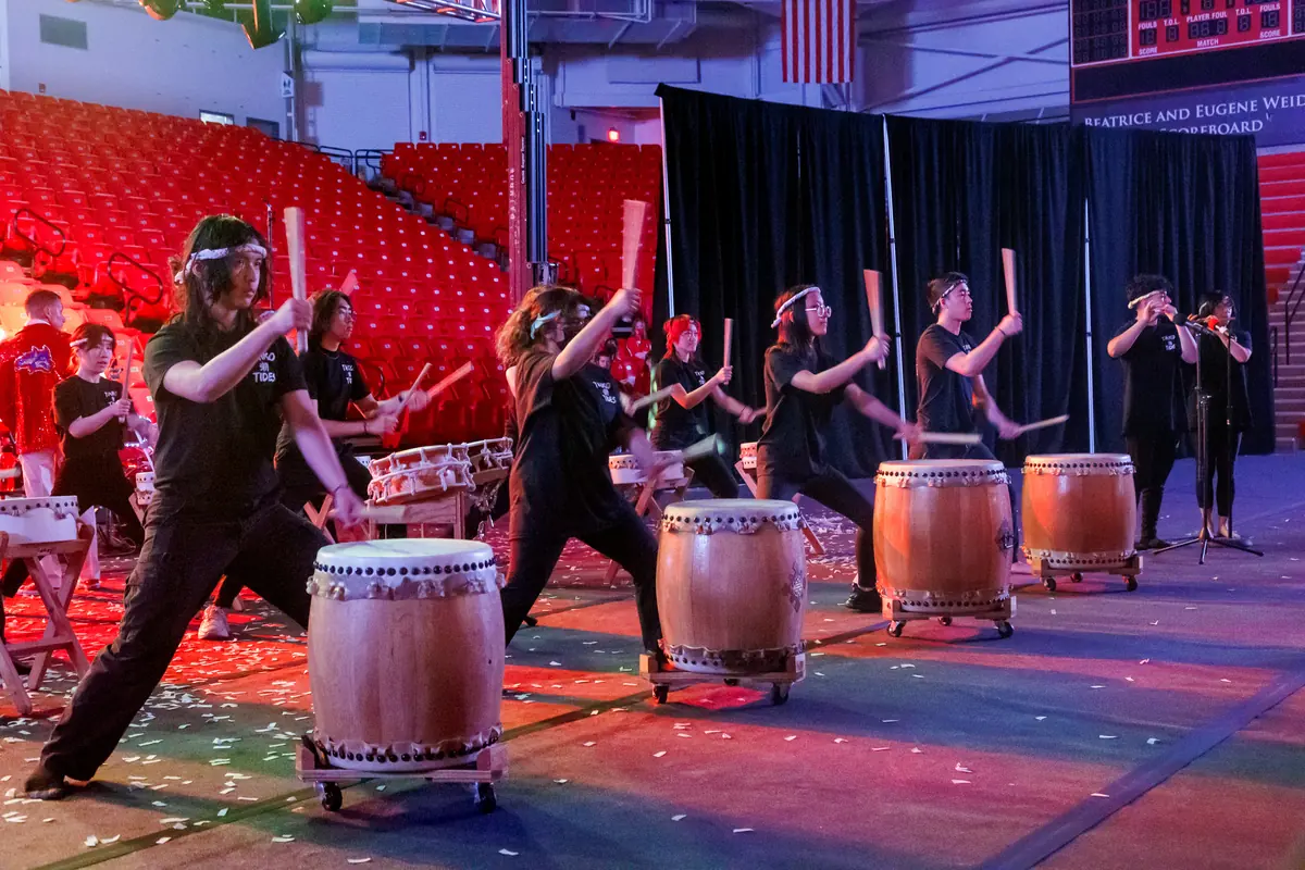 Taiko drummers in black stand behind barrel drums on a stage lit in red and blue, with sticks raised and confetti on the floor