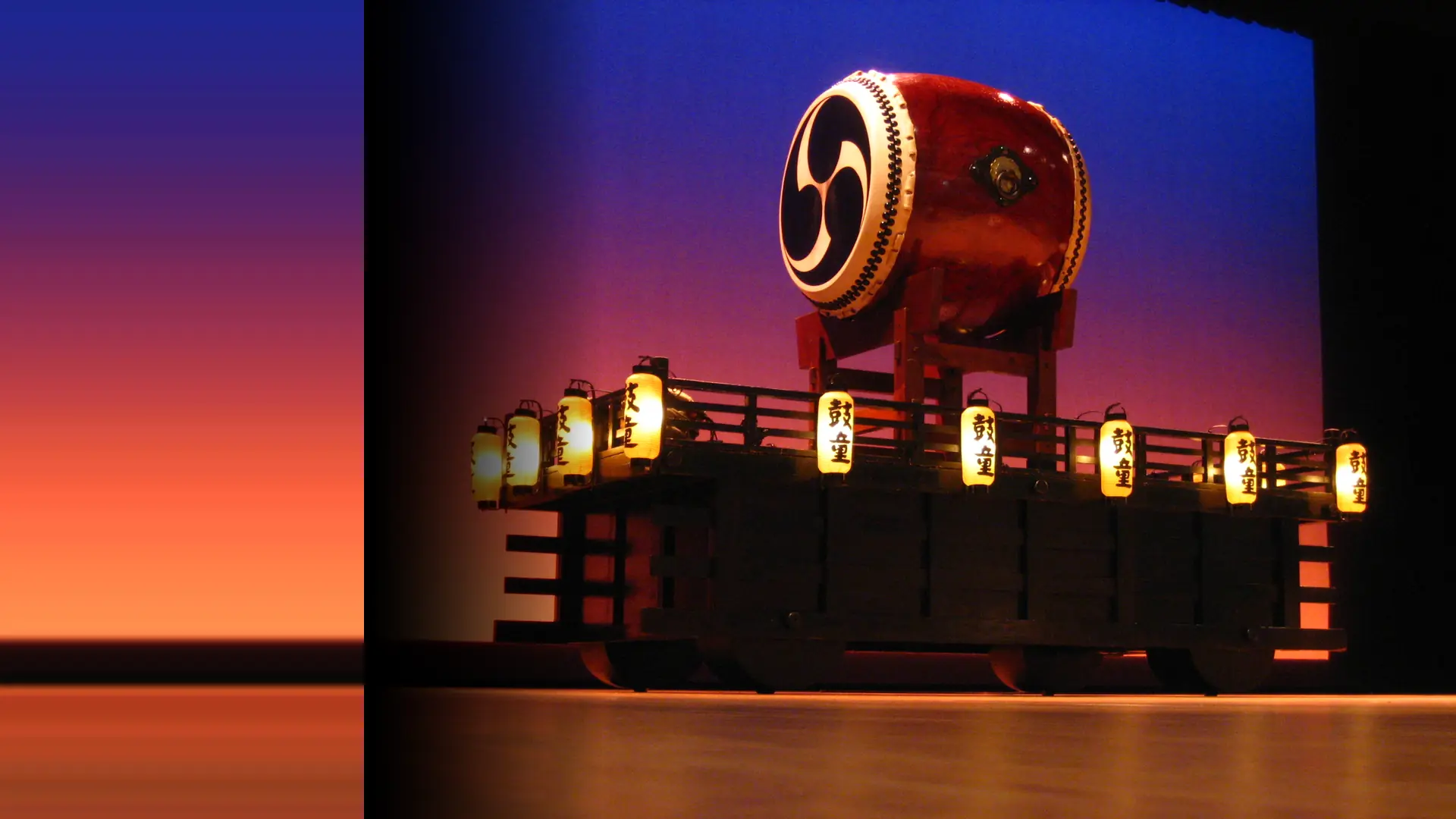 A large red taiko drum with a white and black swirl design sits on a lantern-lit stage platform against a blue and orange backdrop
