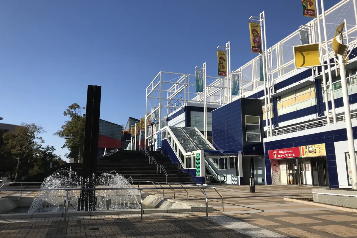 The blue and white Tempozan Marketplace building with banners on tall poles, beside a fountain on a paved plaza under a clear sky